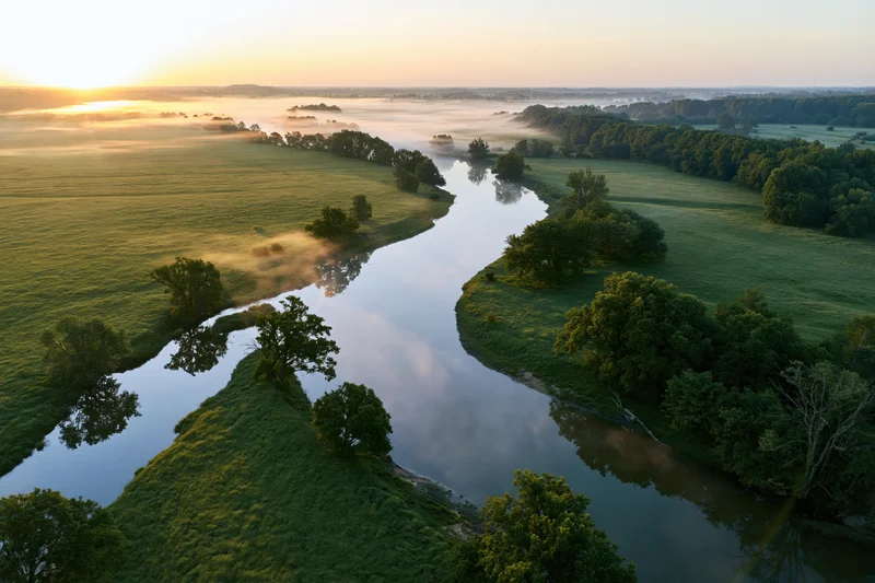 Aerial view of a winding river flowing through green meadows at sunrise with mist over the water and trees along the banks