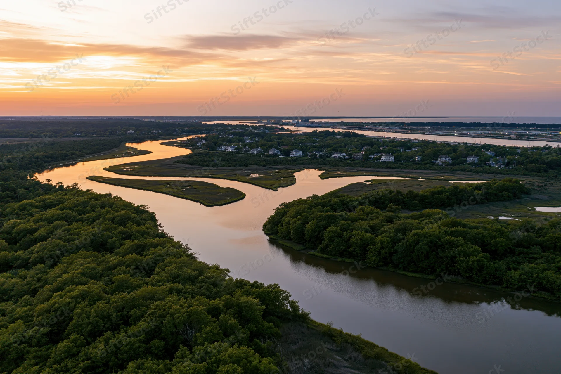 Aerial drone view of a winding tidal marsh estuary at sunset with houses along the riverbank and dense green trees surrounding the water