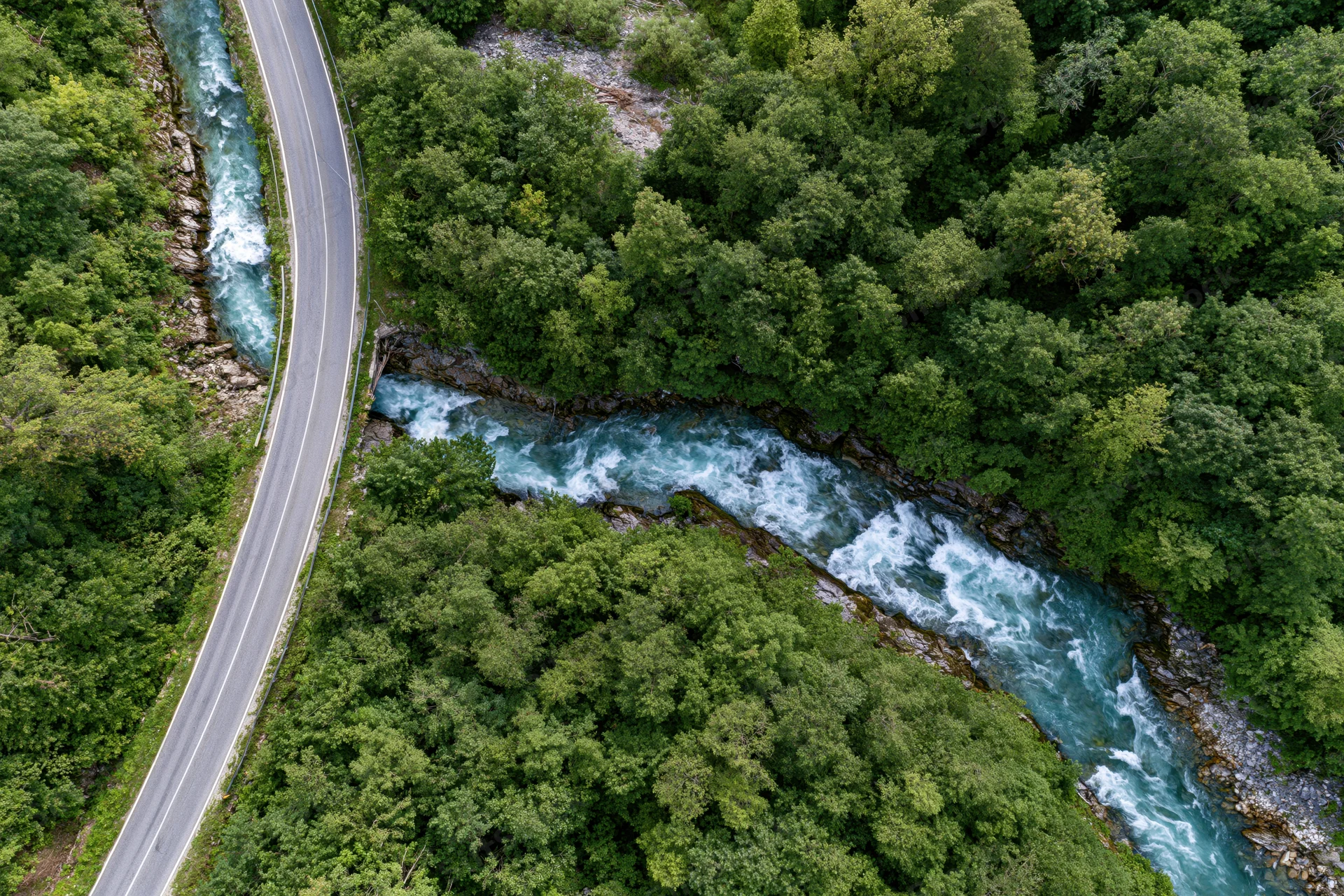 Aerial top-down view of a winding asphalt road alongside a fast-flowing river through dense green forest