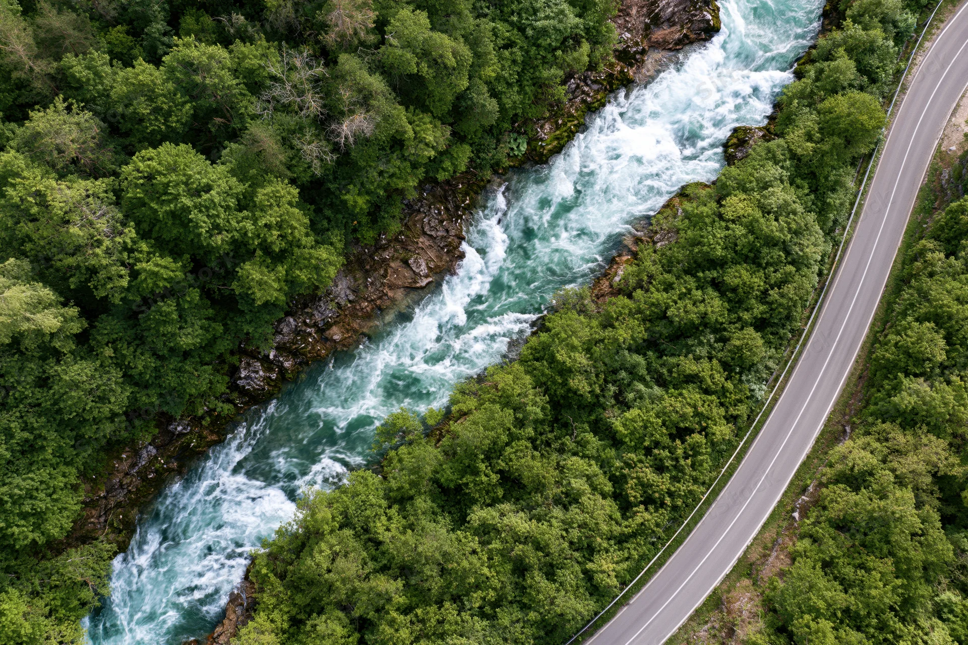 Aerial view of a winding road alongside a fast-flowing river through a dense green forest