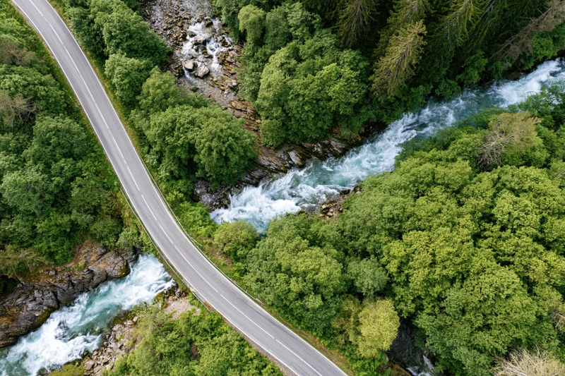 Aerial view of a winding asphalt road alongside a rushing river through a dense green forest