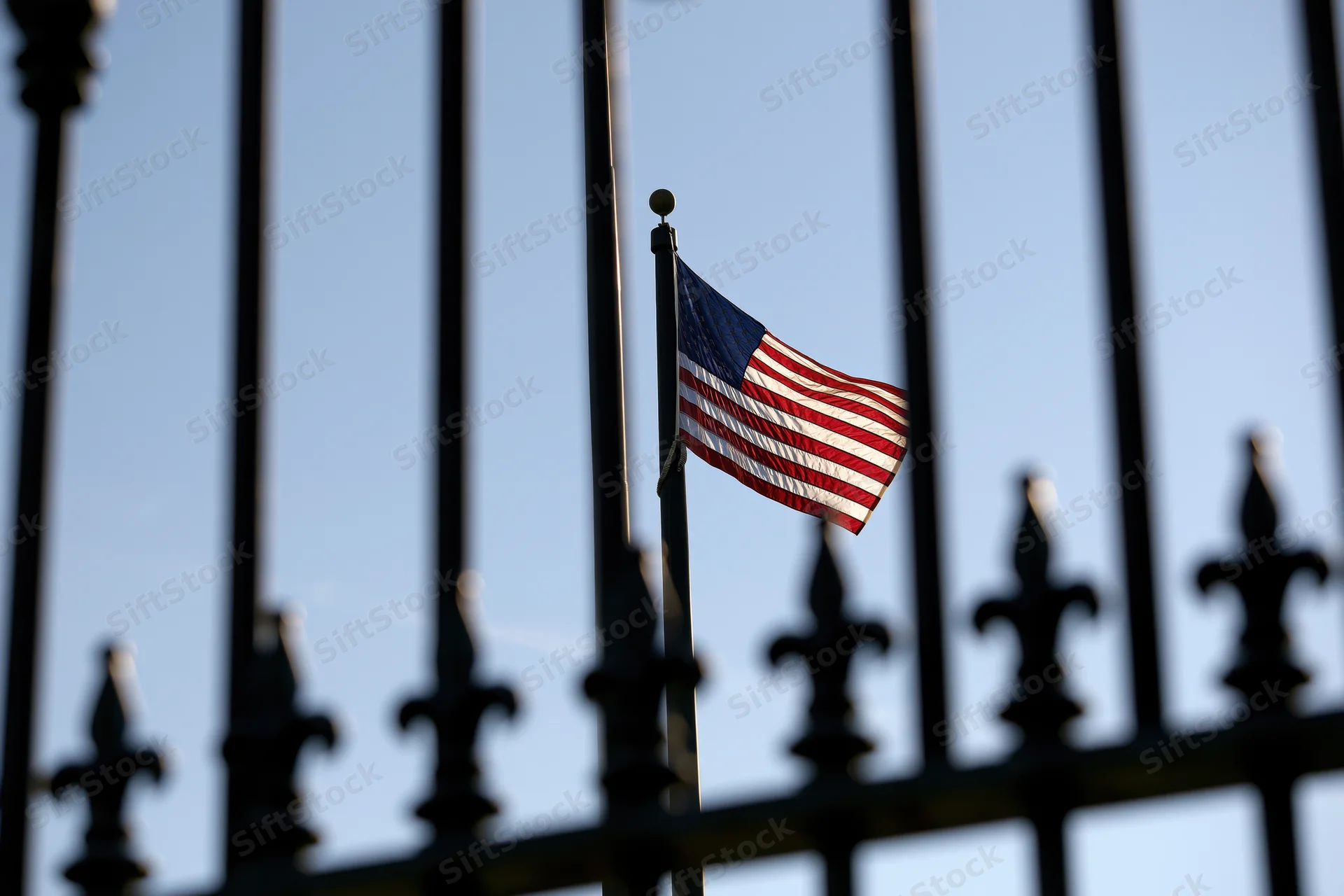 American flag flying on a tall pole behind a wrought iron fence against a clear blue sky