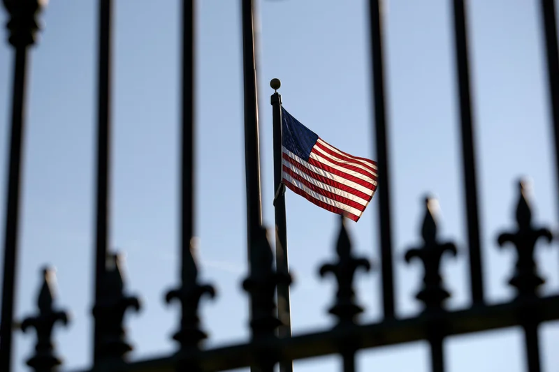 American flag flying on a tall pole behind a wrought iron fence against a clear blue sky