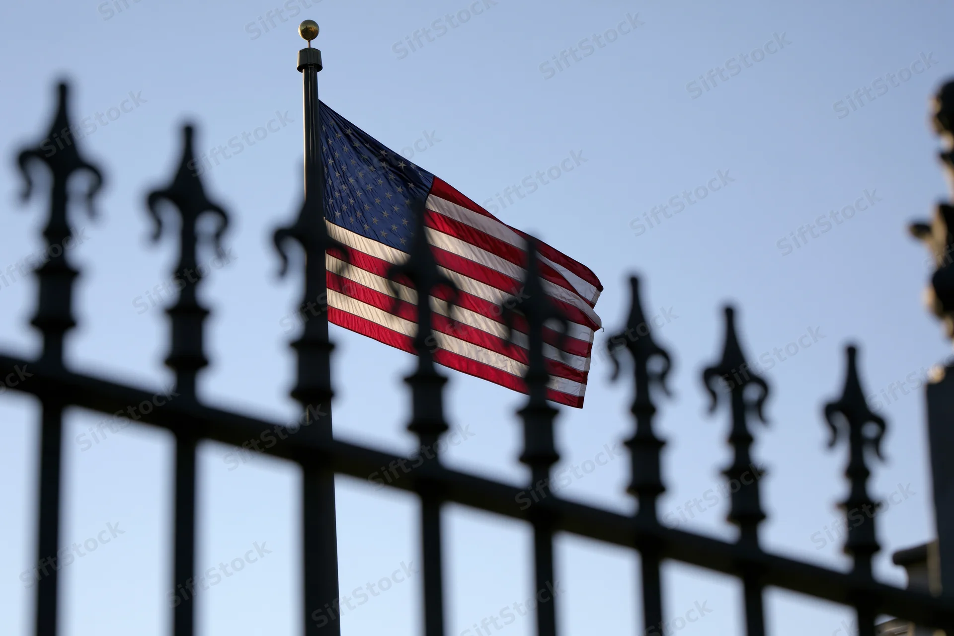 American flag waving on a tall pole behind a black wrought iron fence against a clear blue sky