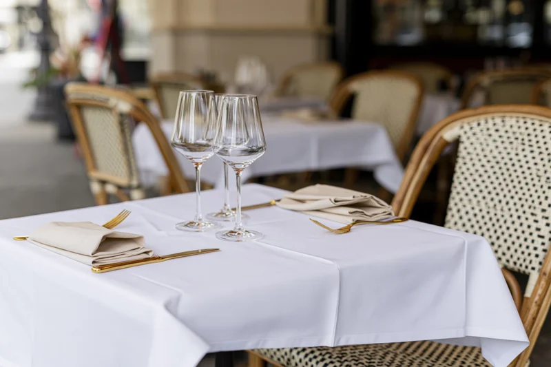 Bistro table set with white tablecloth, wine glasses, folded linen napkins and gold cutlery outdoors