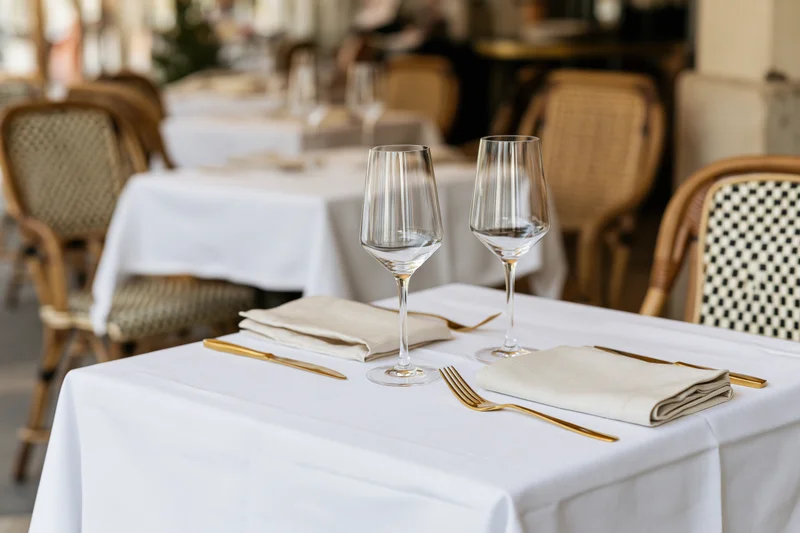 Bistro table set with wine glasses and folded linen napkins on white cloth with rattan chairs in background