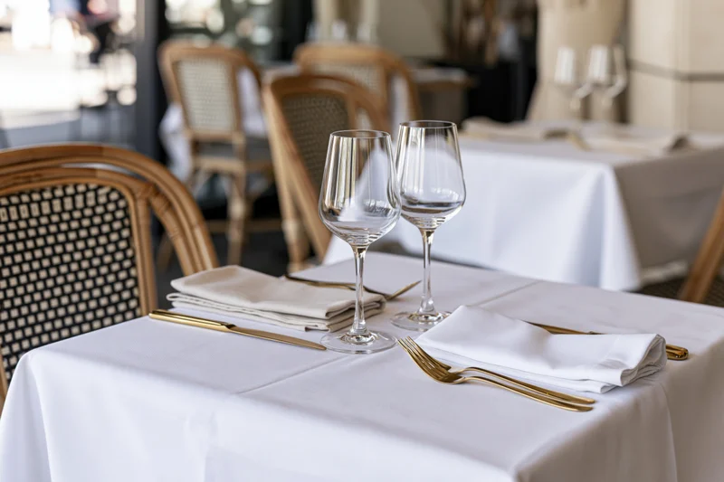 Bistro table set with wine glasses and folded linen napkins on white cloth with gold cutlery