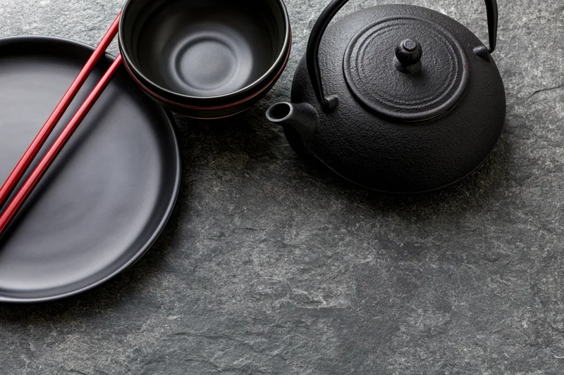 Black cast iron teapot with ceramic bowl and matte black plate holding red chopsticks on stone surface