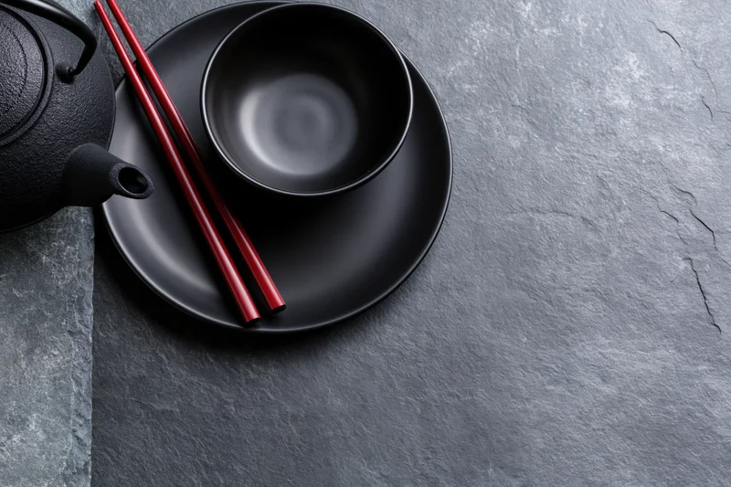 Black cast iron teapot with red chopsticks and matte black bowl on slate surface