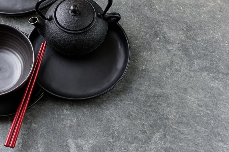 Black cast iron teapot with ceramic bowl and red chopsticks on matte black plate on stone surface