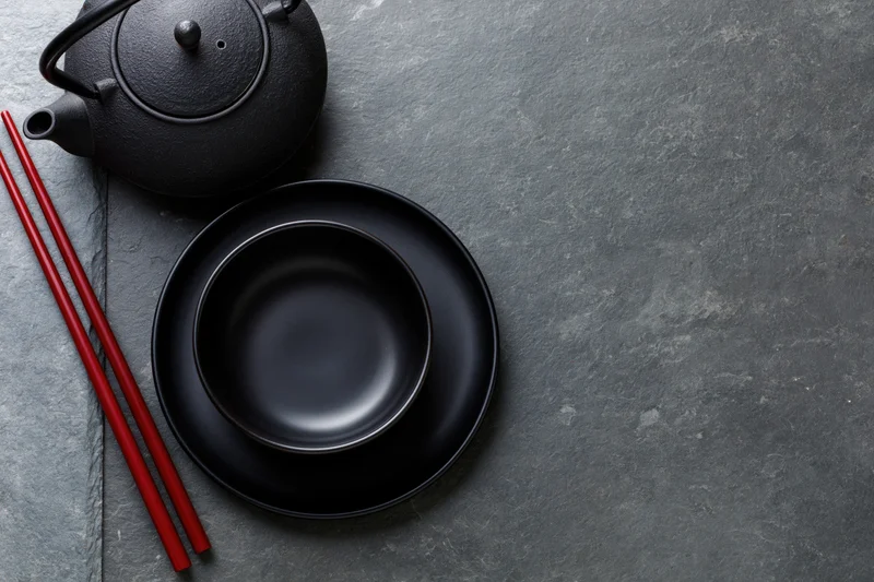 Black cast iron teapot with ceramic bowl and red chopsticks on slate surface