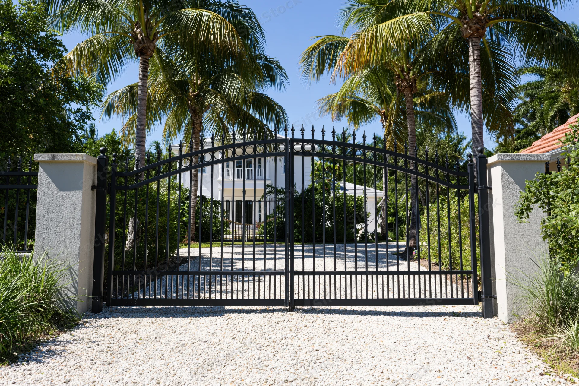 Closed wrought iron arched gate at entrance to white mansion surrounded by palm trees and greenery
