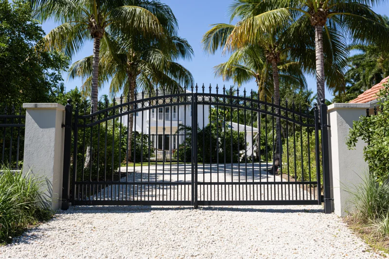 Closed wrought iron arched gate at entrance to white mansion surrounded by palm trees and greenery