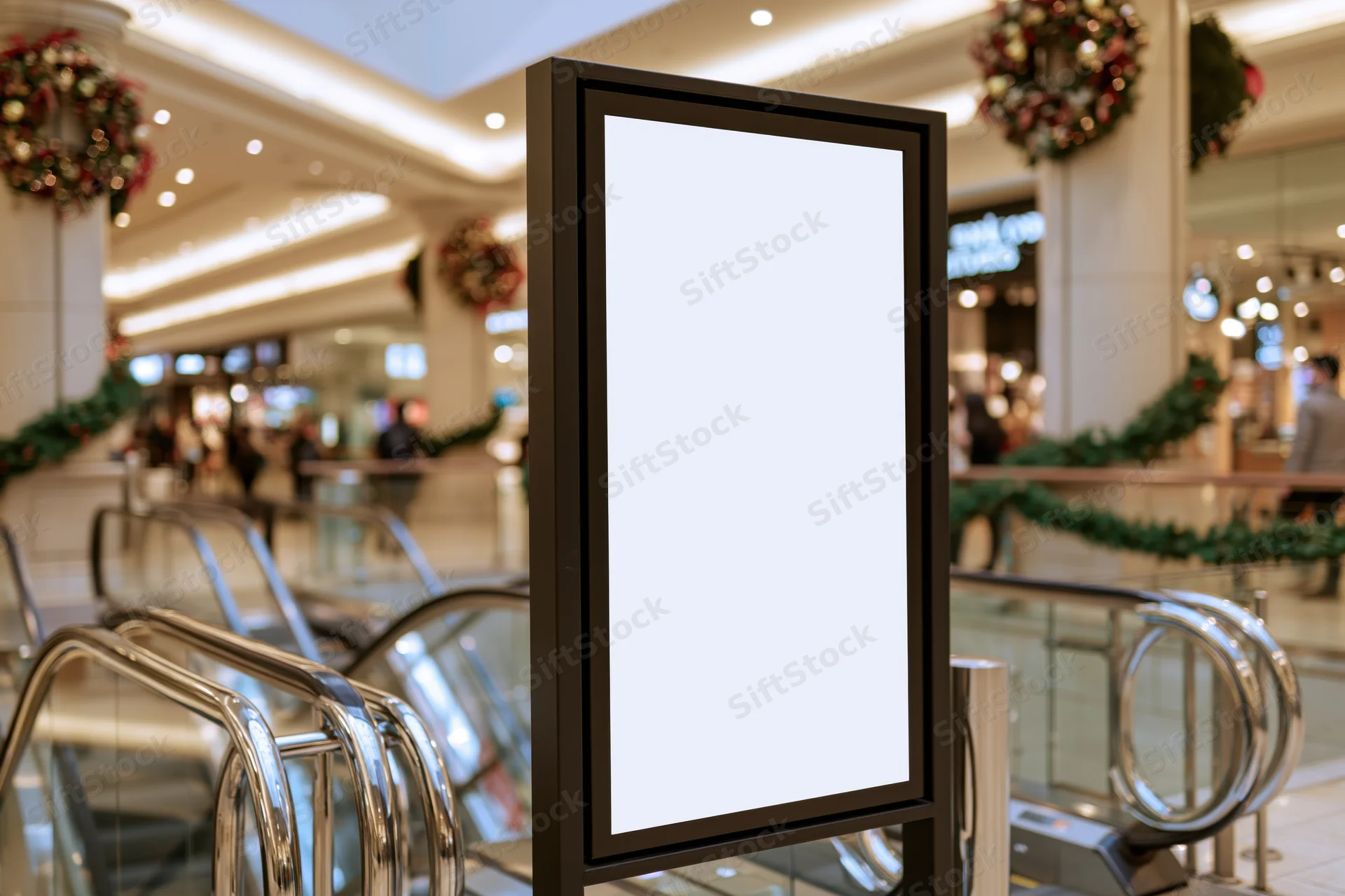 Blank vertical digital display kiosk in a shopping mall with christmas decorations and escalators nearby