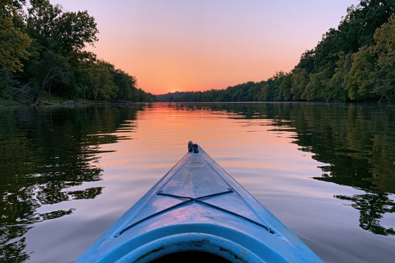 Blue kayak bow gliding down calm river at sunset surrounded by dense trees