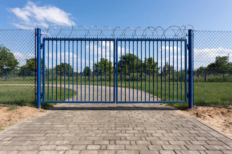 Blue steel security gate with vertical bars and razor wire on chainlink fence at outdoor entrance