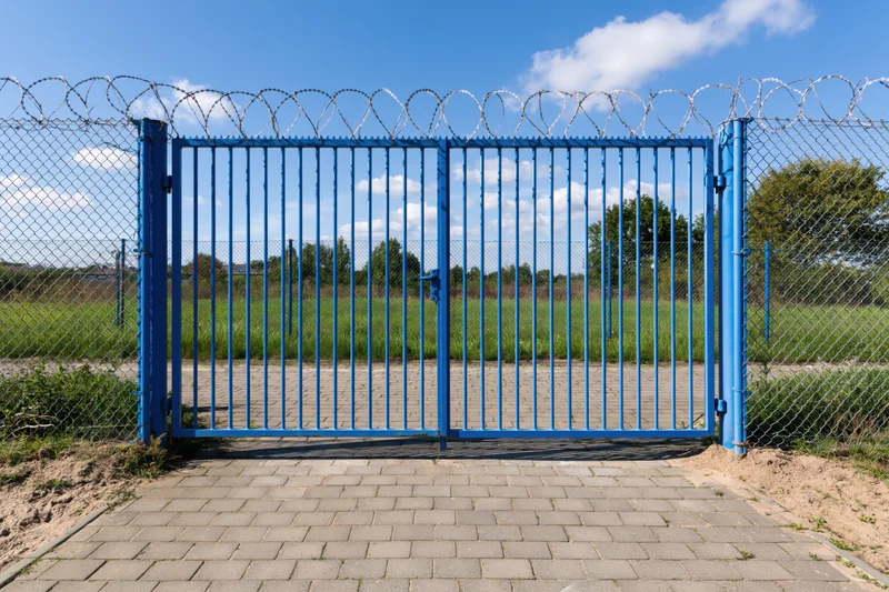 Blue steel security gate with vertical bars and razor wire on chainlink fence at outdoor entrance