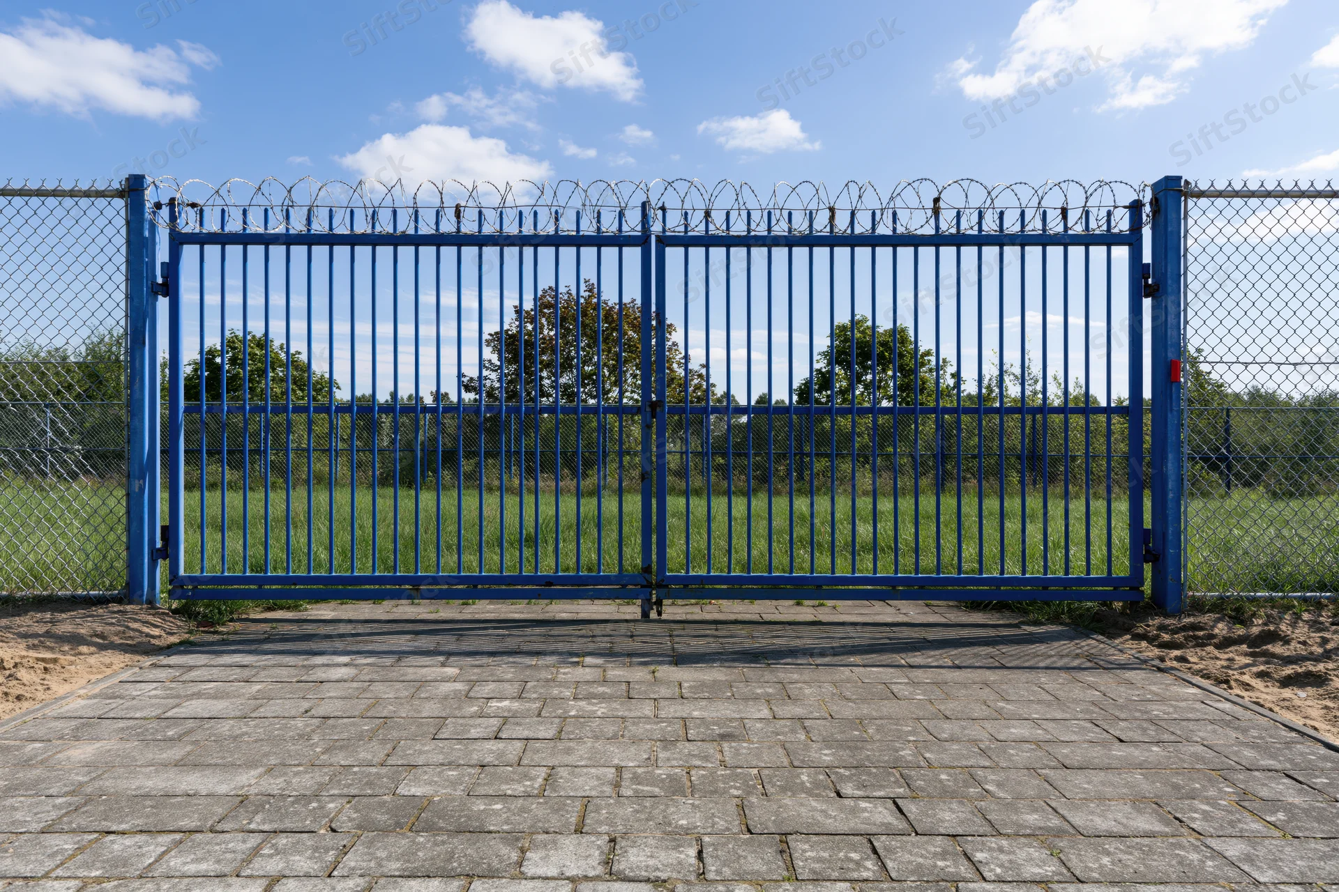 Blue steel security gate with vertical bars and razor wire on chainlink fence, closed, outdoors on paved path leading to grassy field under blue sky