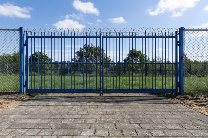 Blue steel security gate with vertical bars and razor wire on chainlink fence, closed, outdoors on paved path leading to grassy field under blue sky
