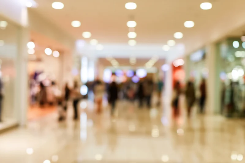 Blurred interior of a busy shopping mall corridor with people walking and storefronts along the sides