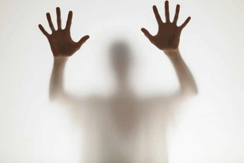 Blurred silhouette of a person pressing hands flat against frosted glass in a bright studio setting