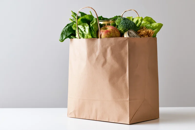 Brown paper grocery bag filled with fresh vegetables and canned goods on white surface