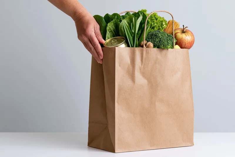 Hand holding a brown paper grocery bag filled with fresh vegetables and groceries on a white surface