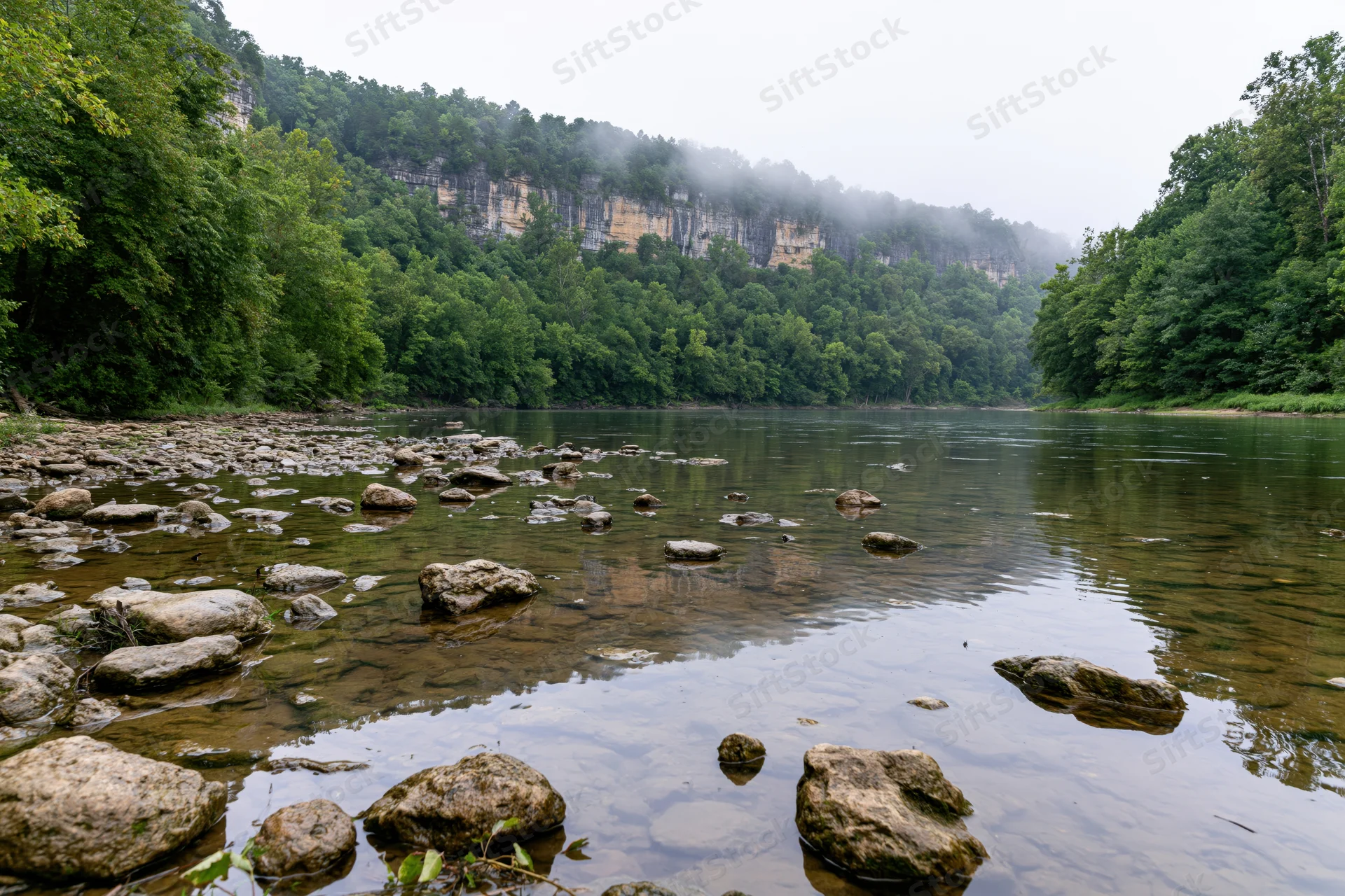 Calm river flowing through limestone bluff canyon surrounded by dense forest and misty sky
