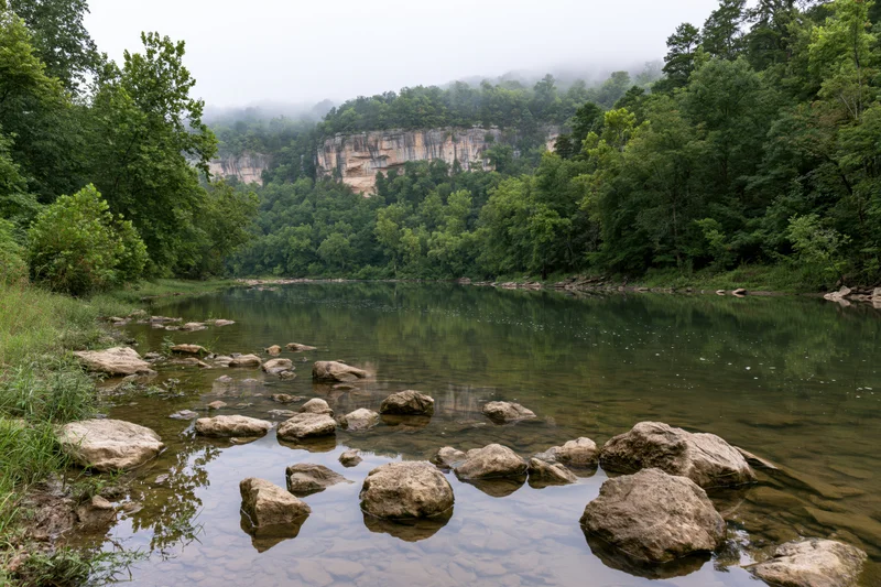 Calm river winding through limestone bluff canyon surrounded by dense green forest under misty overcast sky