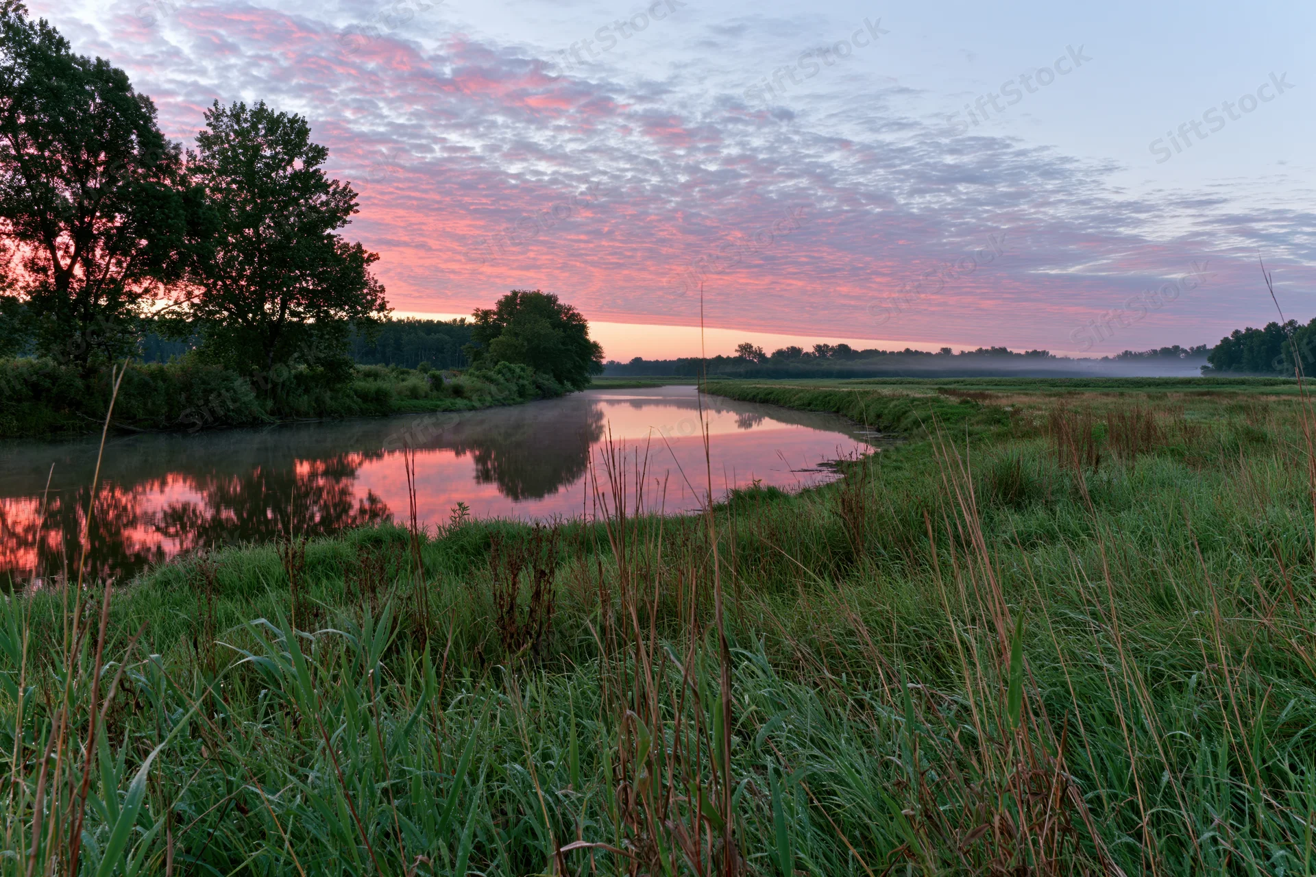 River curving through a green meadow at twilight with pink clouds reflected in the water
