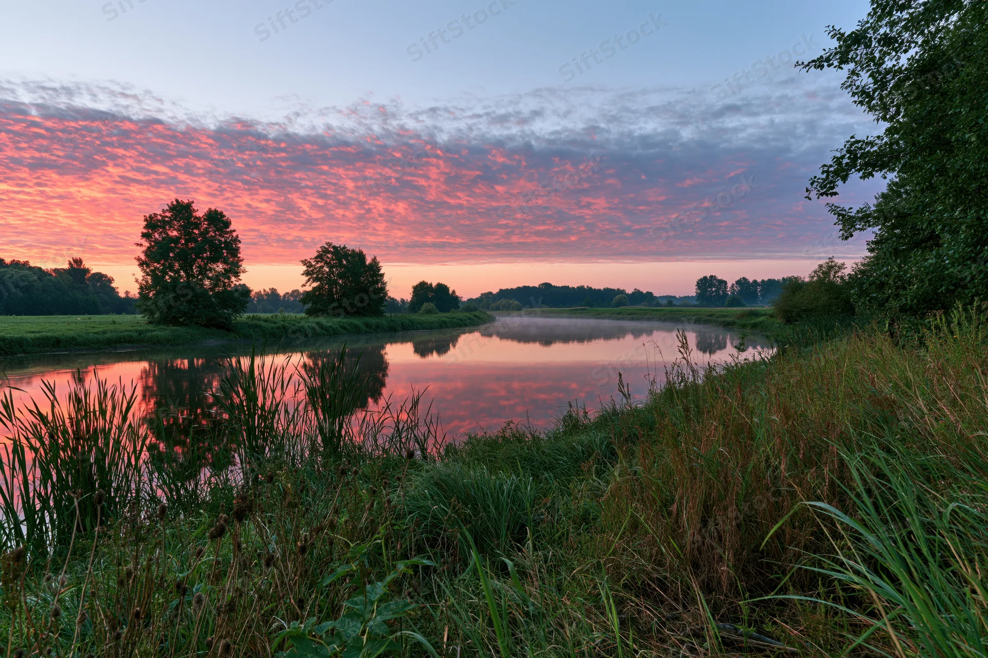Calm river curving through a green meadow at sunset with pink clouds reflected in the water