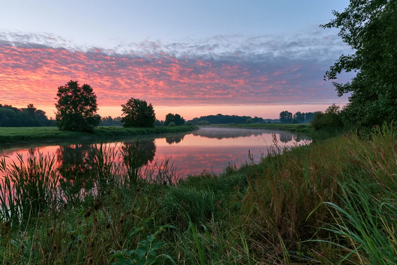 Calm river curving through a green meadow at sunset with pink clouds reflected in the water