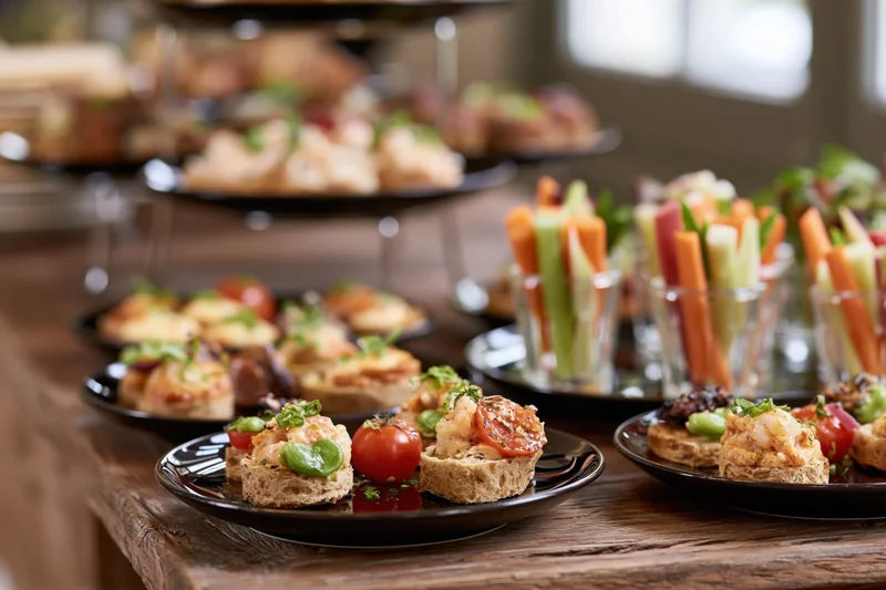 Assorted canapes and vegetable sticks arranged on black plates and glass cups on a wooden table