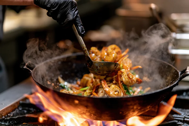 Chef stirring shrimp and vegetables in a cast iron wok over an open flame