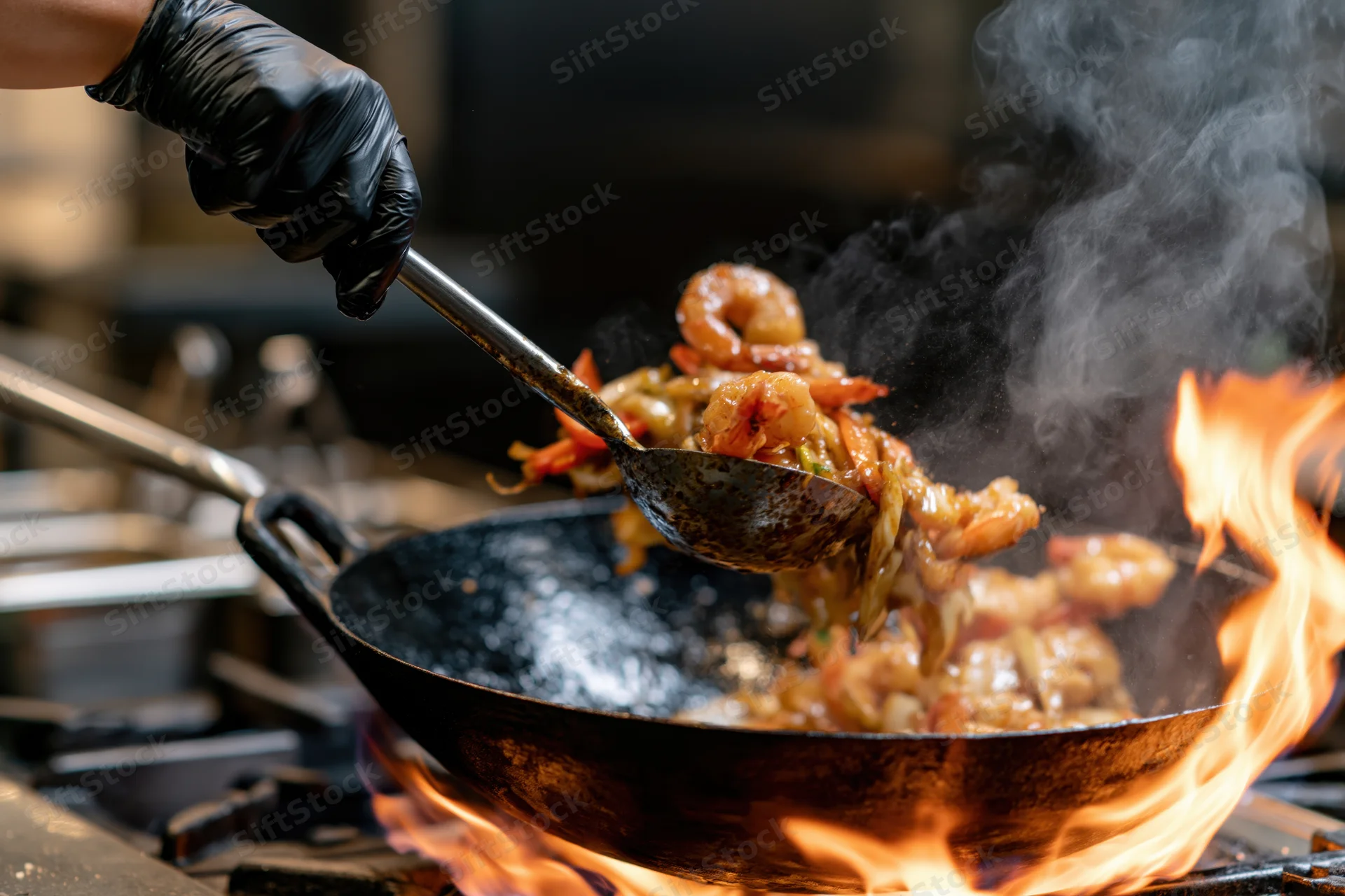 Chef's hand in black glove lifting shrimp from flaming wok with ladle in kitchen