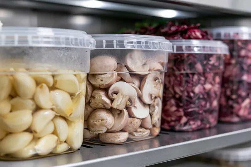 Clear plastic containers holding prepped garlic, mushrooms, and red cabbage on a stainless steel shelf in a refrigerator