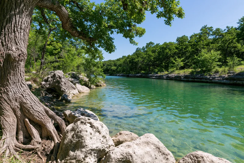Clear turquoise river flowing through limestone banks surrounded by green trees and rocks under blue sky