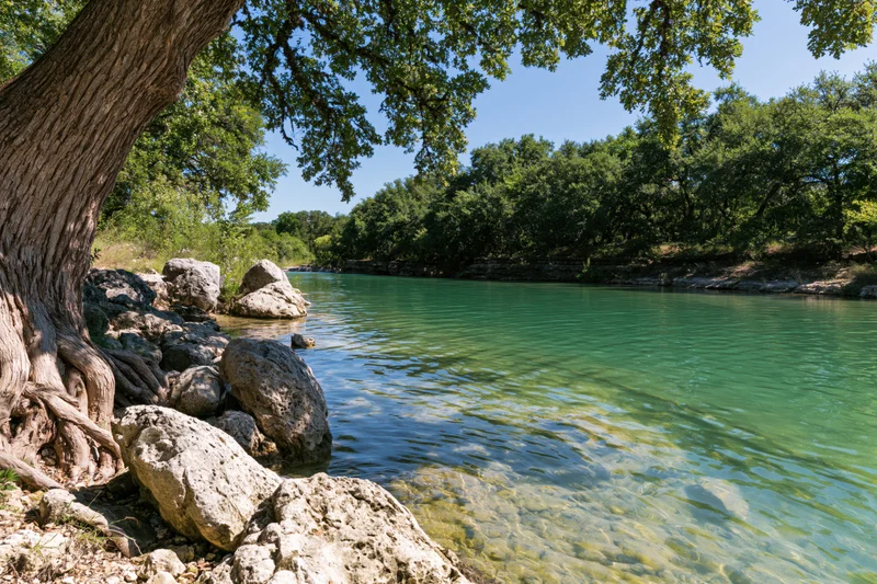 Clear turquoise river flowing beside limestone banks and oak trees under a blue sky