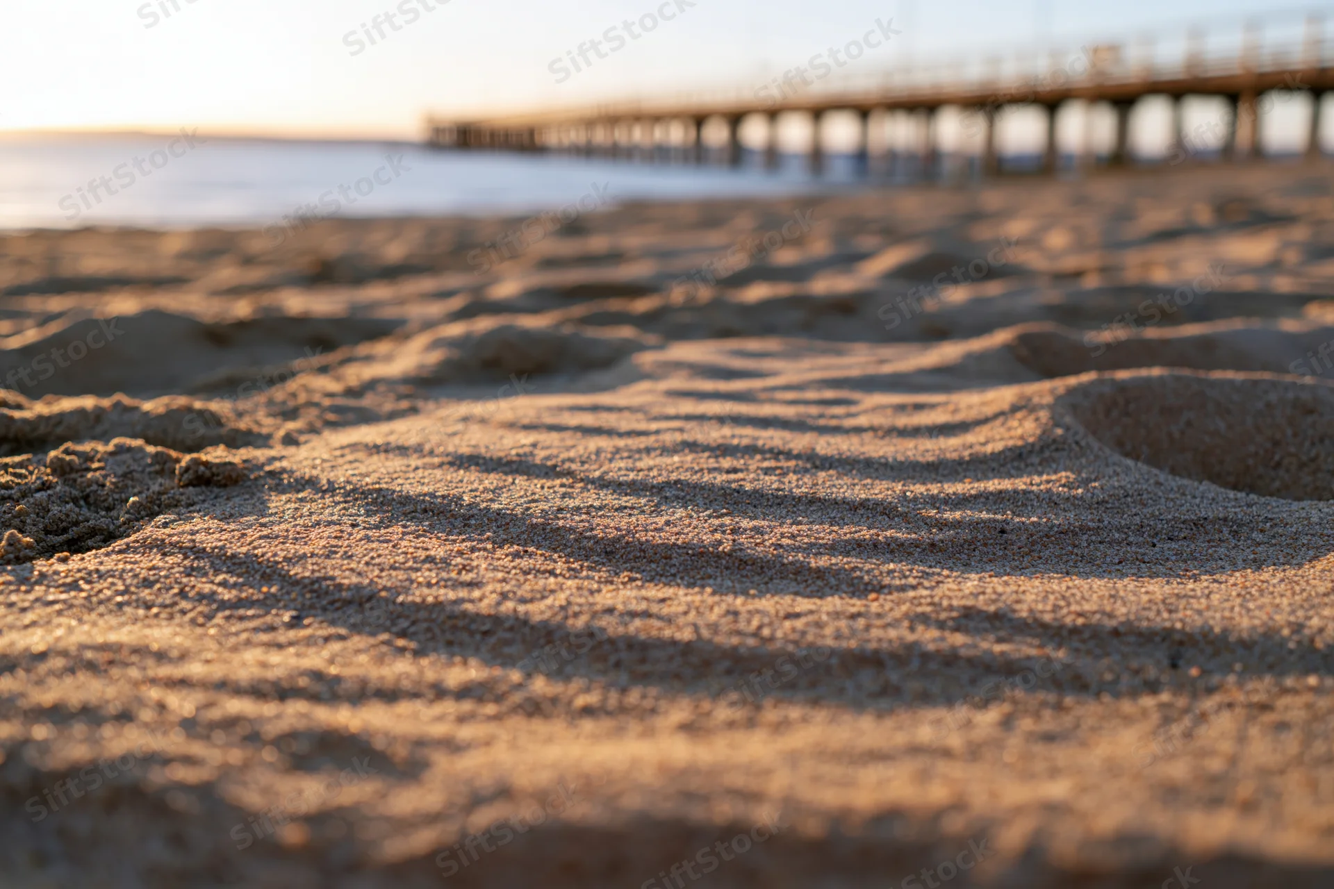 Close-up of rippled dry sand on a beach at sunset with a wooden pier in the distance