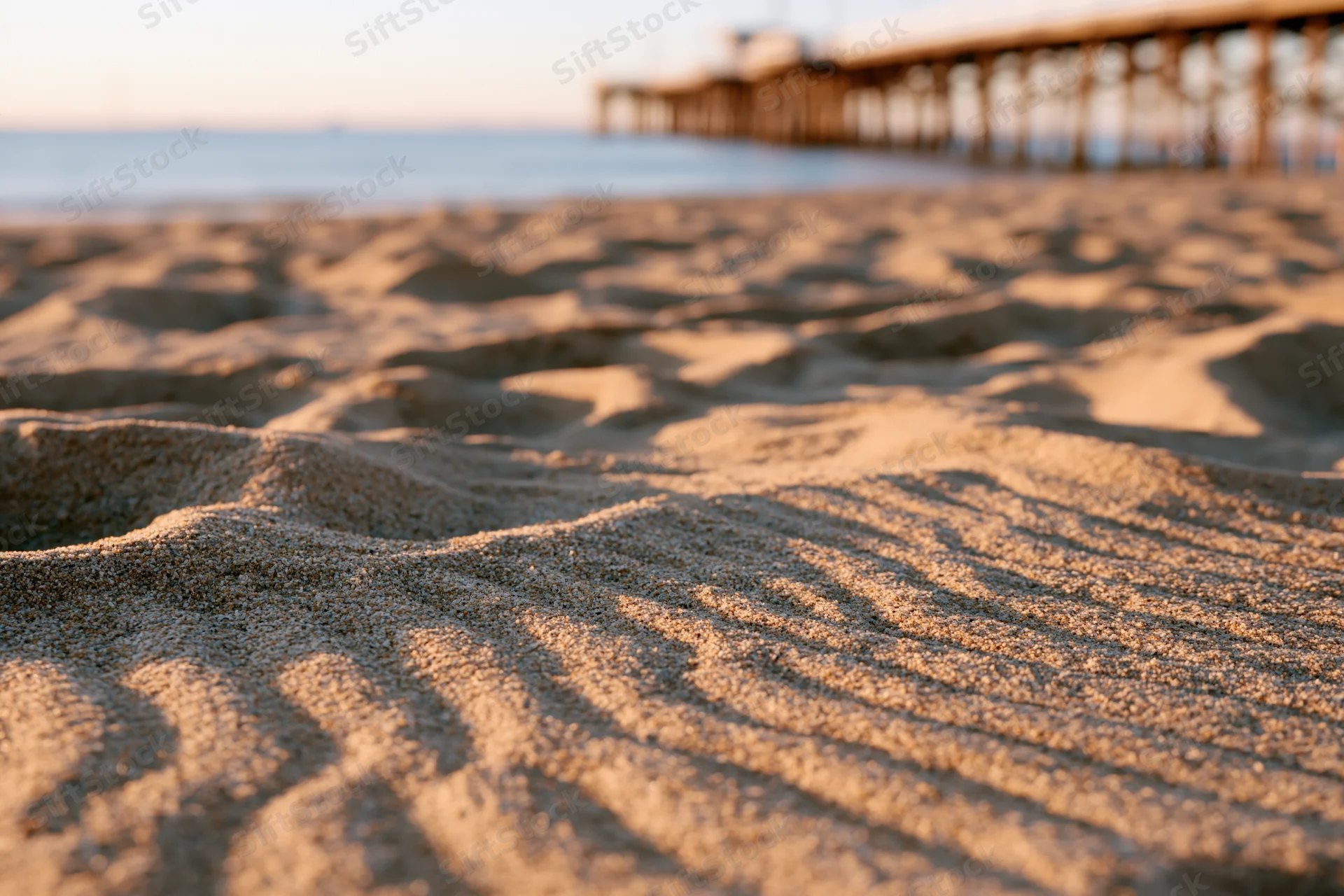 Closeup of rippled dry beach sand with wooden pier in blurred background at sunset