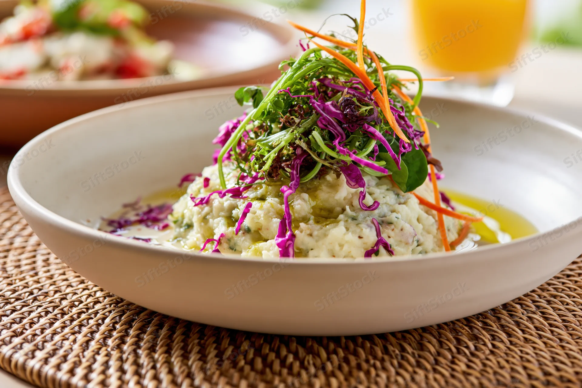 Close-up of a mound of creamy mashed potato dish topped with microgreens and shredded vegetables in a white bowl on a woven placemat