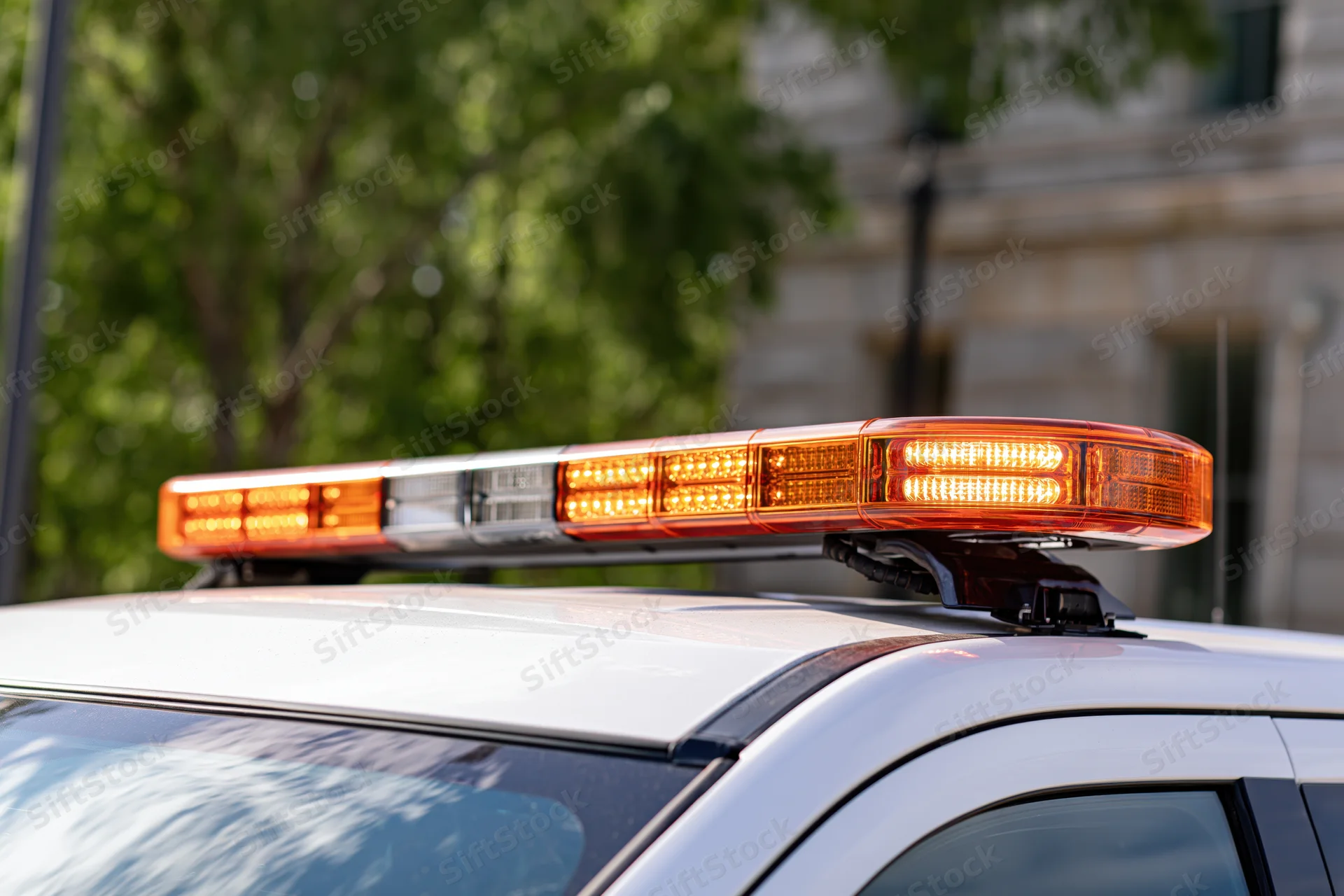 Amber and white led light bar mounted on top of a police vehicle roof with trees and building in background