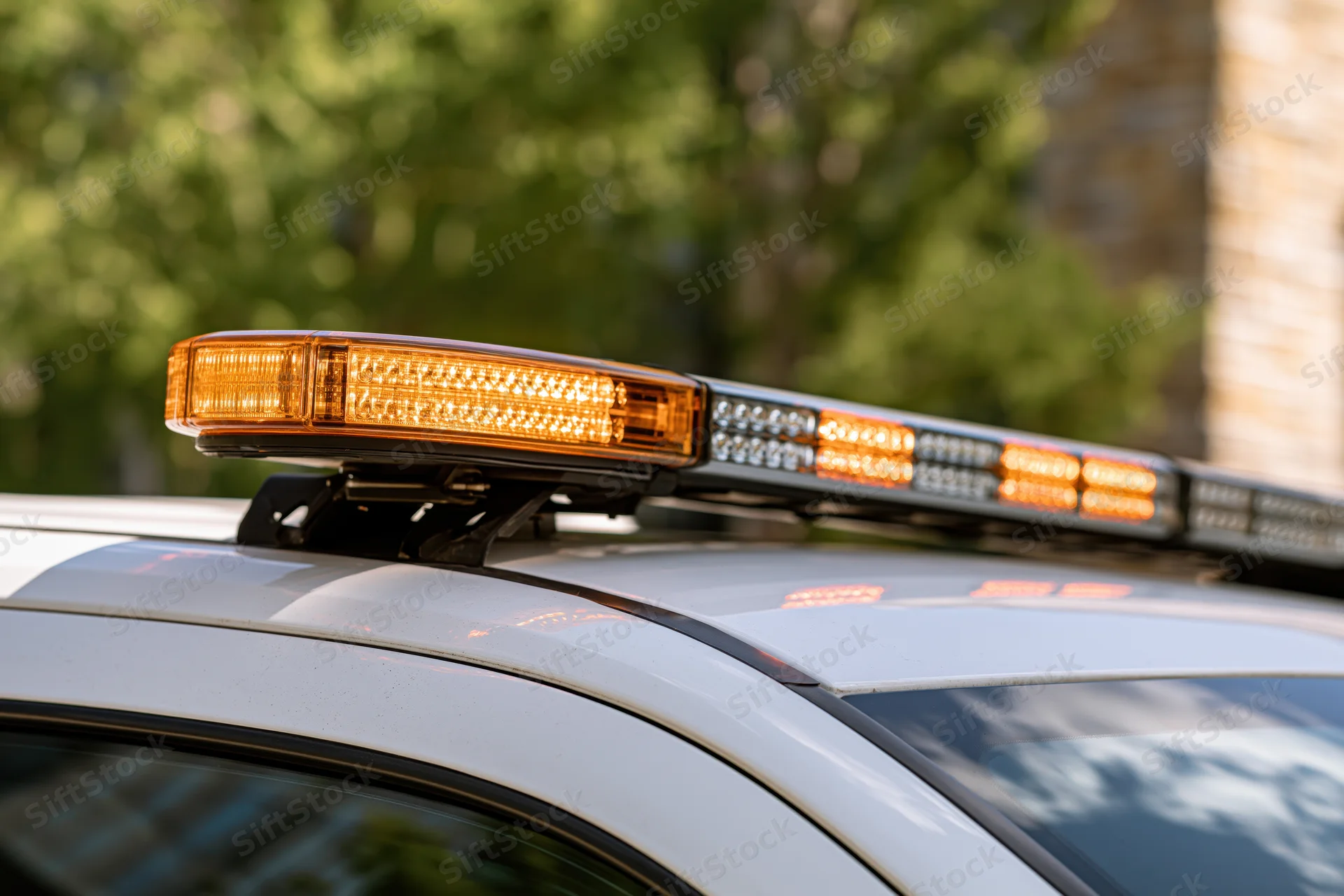 Amber and white led light bar mounted on top of a white police vehicle roof with trees in background
