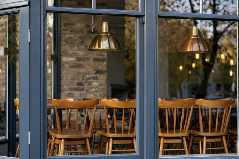 Restaurant interior with wooden chairs and brass pendant lights viewed through large glass windows from the street