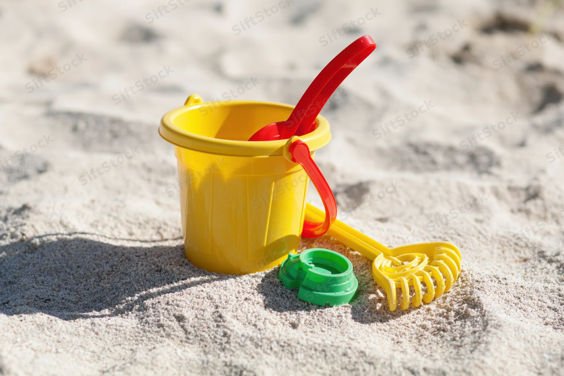 Colorful plastic children's beach toys including yellow bucket, red shovel, and green mold on sandy shore