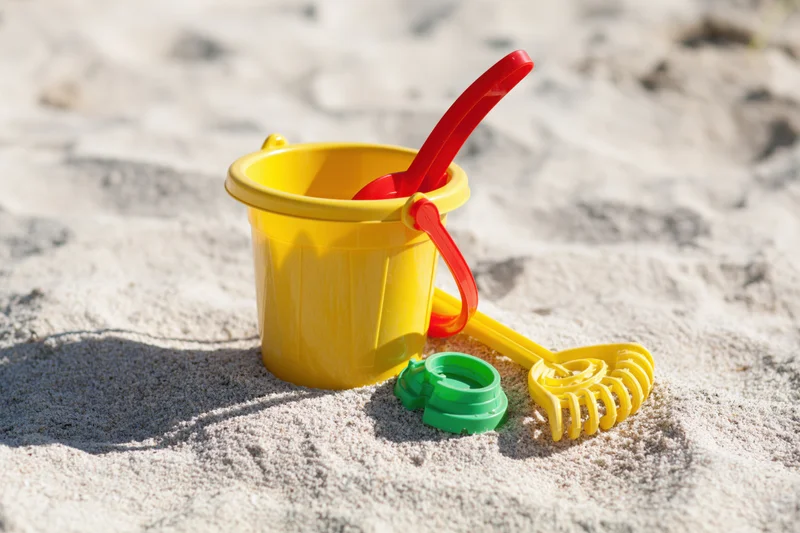 Colorful plastic children's beach toys including yellow bucket, red shovel, and green mold on sandy shore