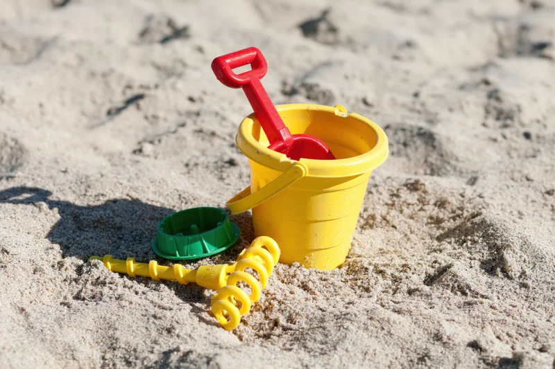 Colorful plastic children's beach toys including yellow bucket, red shovel, green mold and yellow spiral tool resting in sand