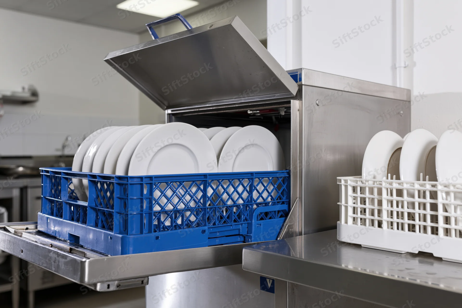 Commercial dishwasher with open hood loading white plates into blue crate in kitchen