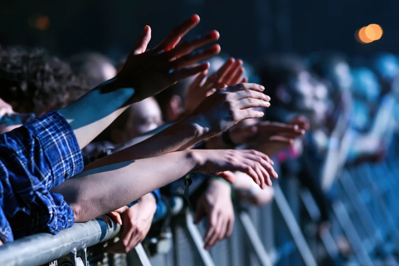 Concert crowd reaching arms over metal barrier toward stage at night