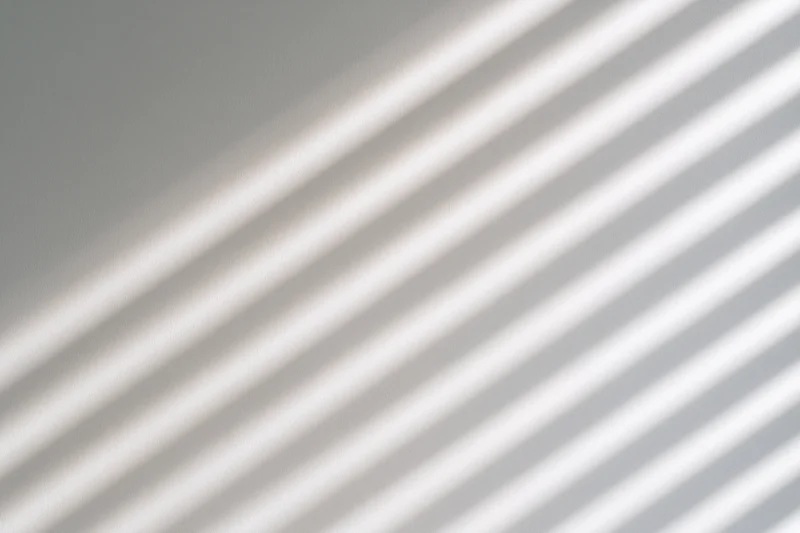 Diagonal shadows from window blinds cast across a plain white wall in a bright interior
